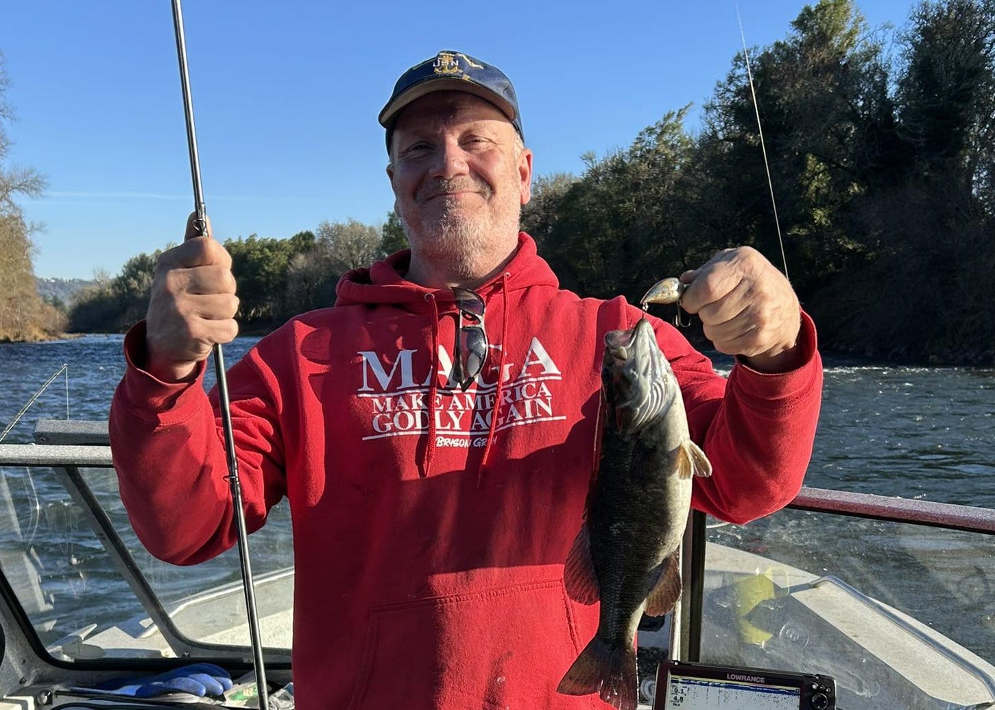 Facebook profile of Charles Simmons, a middle-aged man with grey stubbly beard, blue US Navy baseball cap, and red 'Make America Godly Again' sweatshirt; he stands in a powerboat on a river, displaying a fishing rod and a bass he's just caught and lectured about how taxation is theft. He captioned the photo ‘This one won’t be eating salmon smolts!’ as if he’d done a public service.