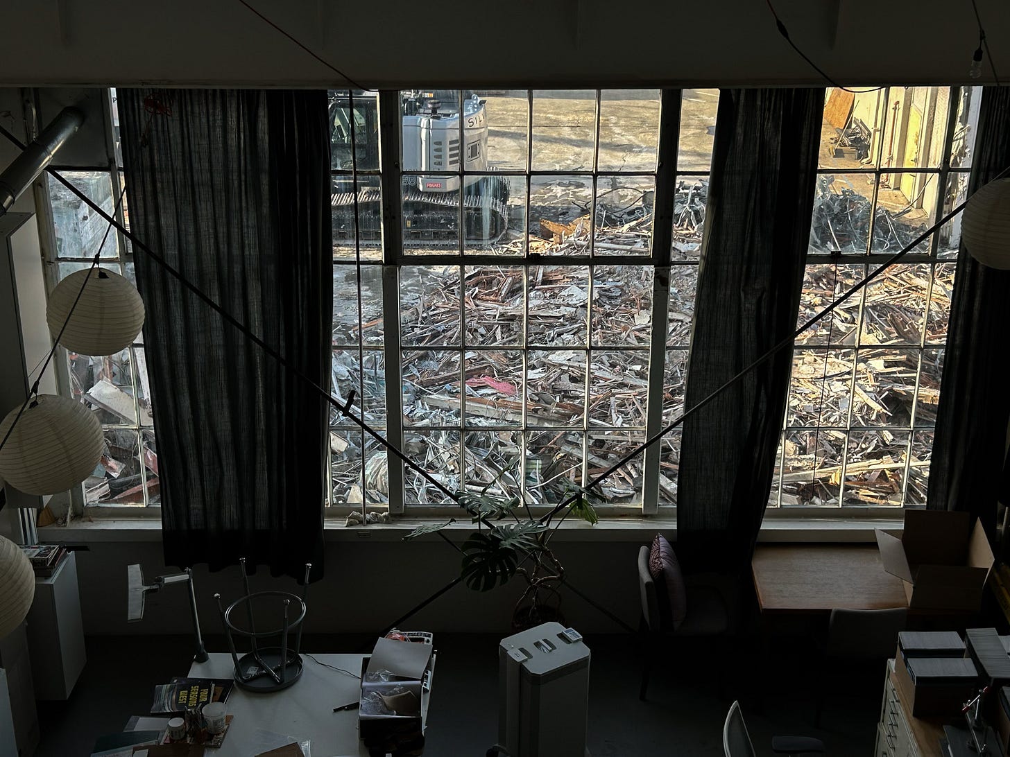 A studio interior is shown in partial shadow, with a view through large industrial-style grid windows revealing a demolition site outside. The exterior is filled with rubble, splintered wood, and twisted metal debris from a recently collapsed building. Part of a Silverado-branded excavator is visible near the top left of the window frame. Inside the studio, there are tables, chairs, an air filter, a potted Monstera plant, hanging paper lanterns, and various scattered publications and artworks in-progress. Dark curtains are partially drawn to the sides. The scene captures a stark contrast between the active destruction outside and the quiet workspace within.
