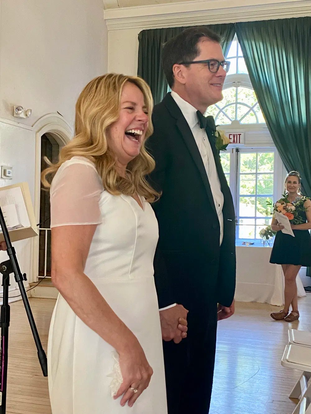 Blonde bride and brown haired groom with glasses, holding hands. Windows with green curtains behind the couple.