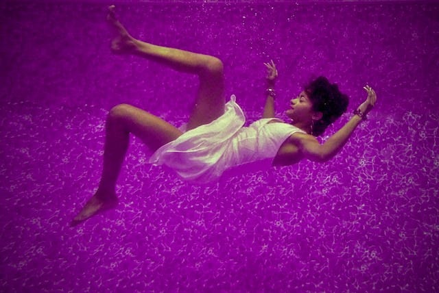 A woman submerged underwater at the bottom of a pool, lying horizontally with a serene expression, her eyes closed in a dreamlike state, symbolizing hope and longing.