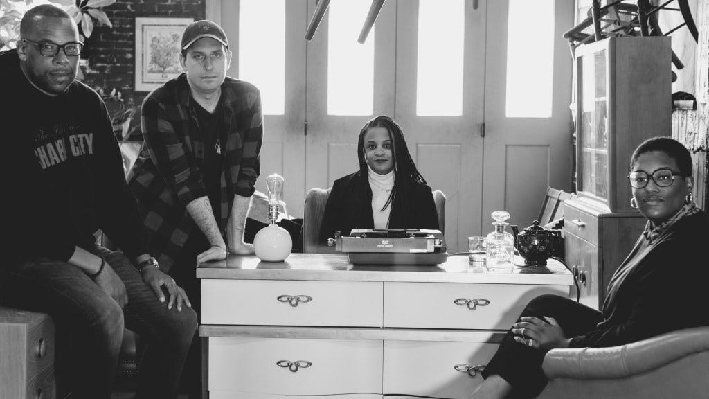Photo of the Beat Staff, in black and white, photographed by Schaun Champion. Left to right:  J. Brian Charles sitting, Brandon Soderberg leaning forward, Lisa Snowden sitting at a desk, and Teri Henderson sitting in a chair.