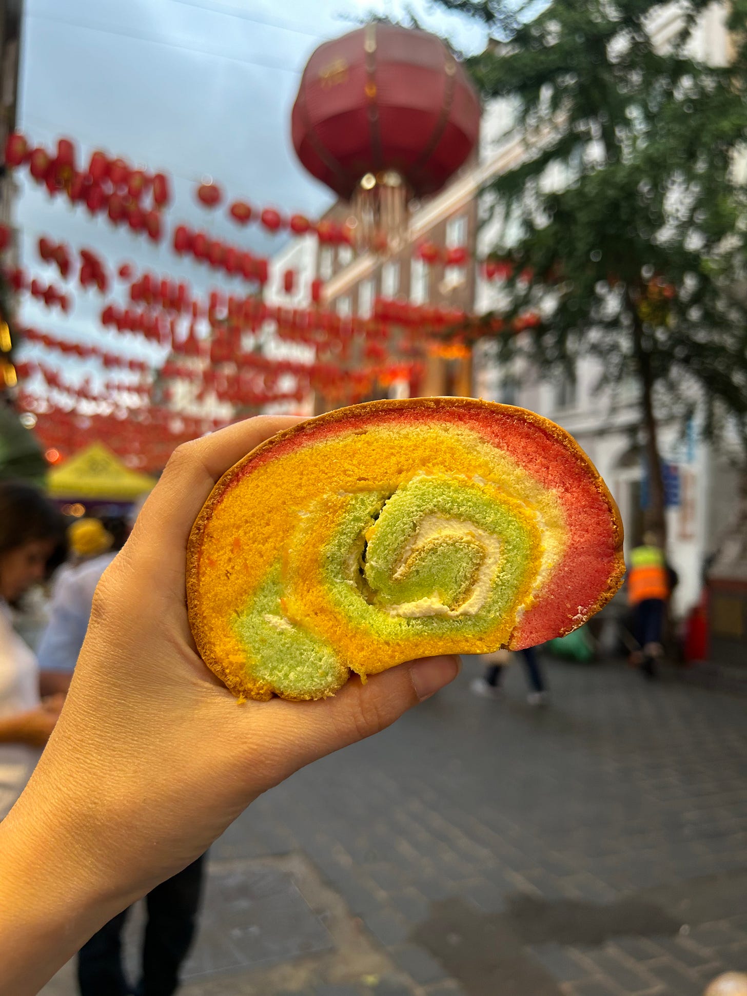 A hand holds up a Swiss roll marbled in pink, orange, and green. There are lanterns strewn across buildings in the background.