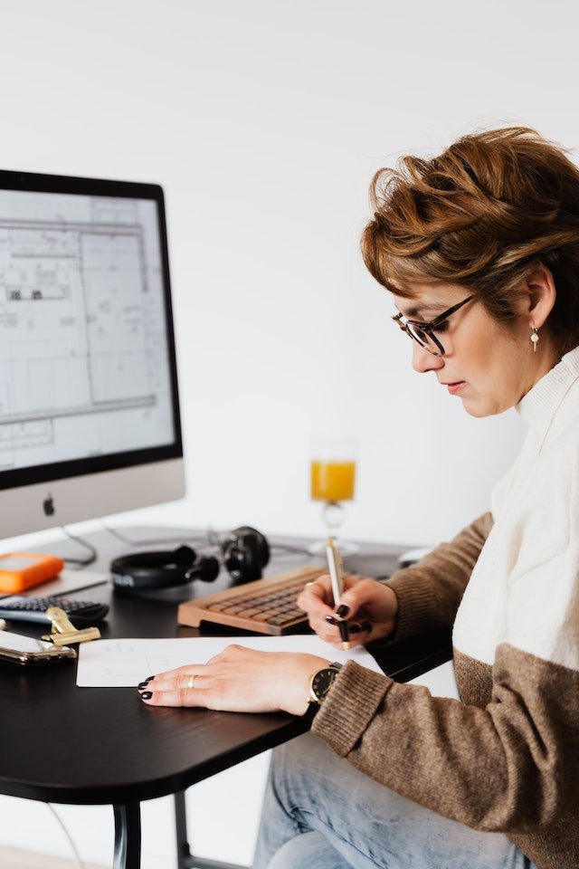 A woman sitting in front of a desktop computer, but turned to the side and writing something down on a piece of paper with a pen.