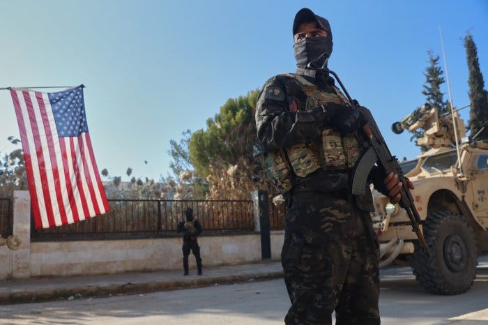 A fighter with the Kurdish-led Syrian Democratic Forces stands guard as an American flag hangs in the background. A fighter with the Kurdish-led Syrian Democratic Forces stands guard as an American flag hangs in the background.