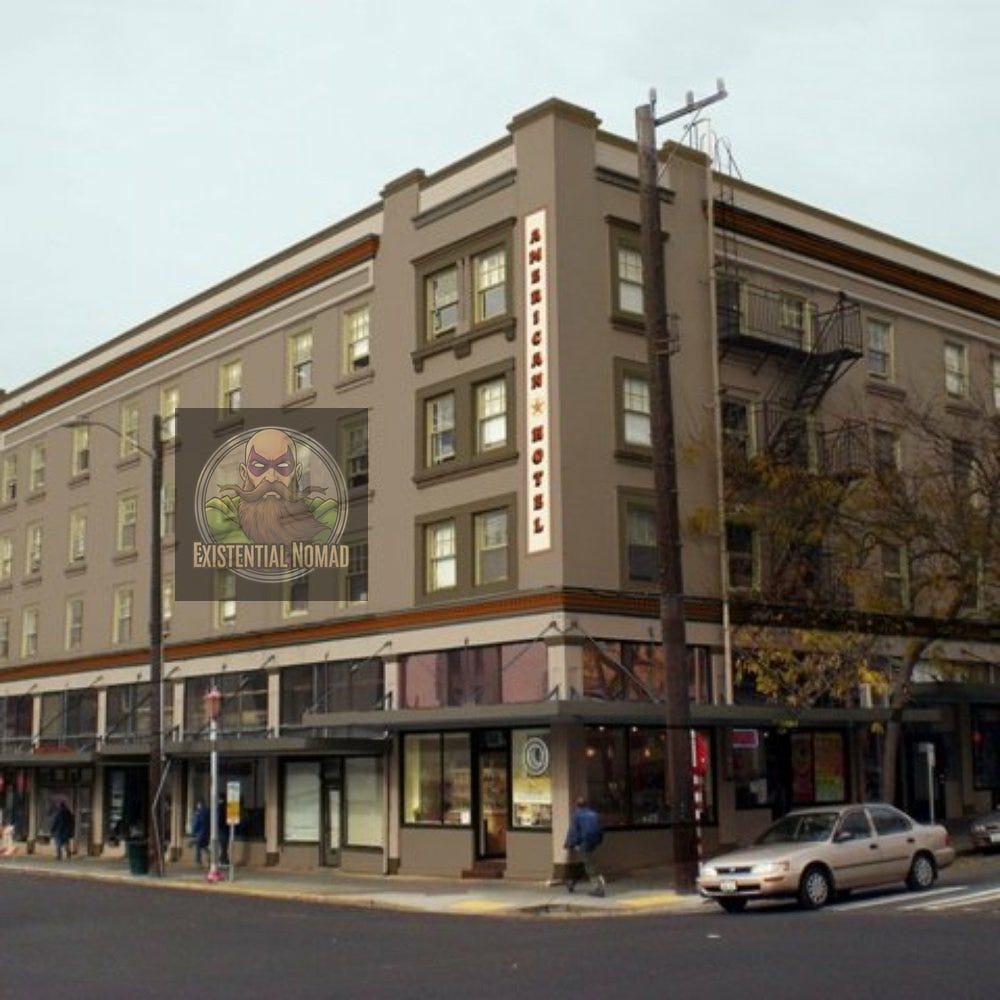  This is a photograph of a multi-story building on a city corner. A vertical sign on the corner of the building reads "AMERICAN HOSTEL" with a star. The first floor has large windows for commercial use, while the upper floors have rows of smaller windows. The building is painted a neutral color with a darker trim. A street sign and power pole are visible on the corner, and a car and several pedestrians are on the street below.