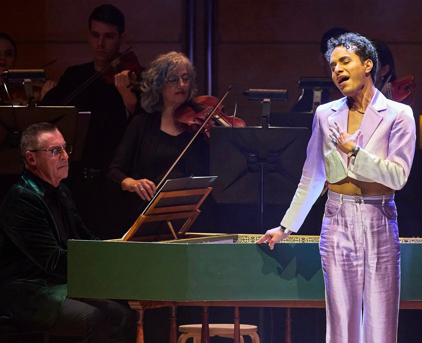 Opera singer Samuel Mariño wearing a lilac pantsuit and standing singing alongside a man playing a green harpsichord. Opera singer Samuel Mariño wearing a lilac pantsuit and standing singing alongside a man playing a green harpsichord.