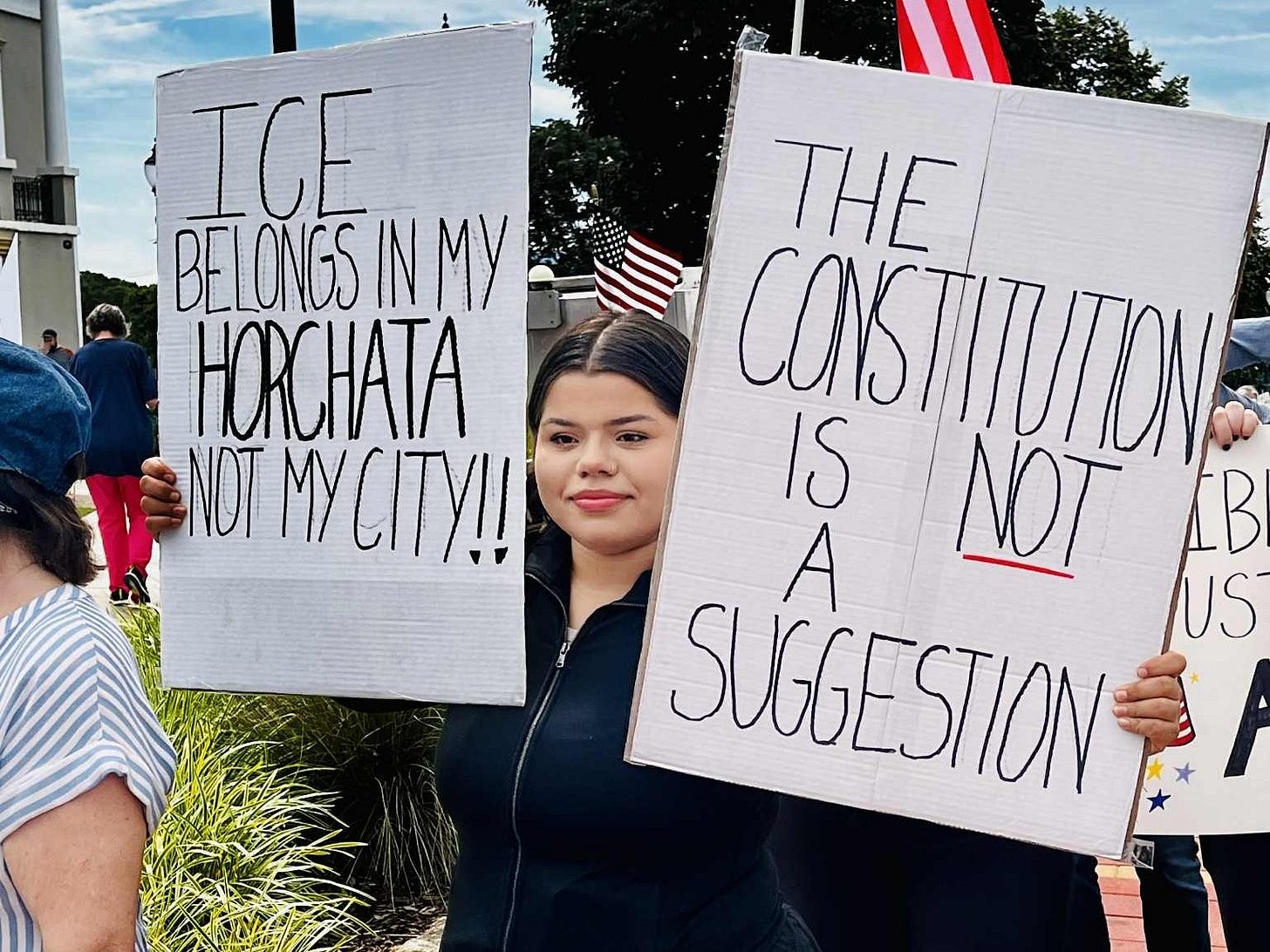 A protester holds two signs, one reading "ICE belongs in my horchtata not my city!!"
