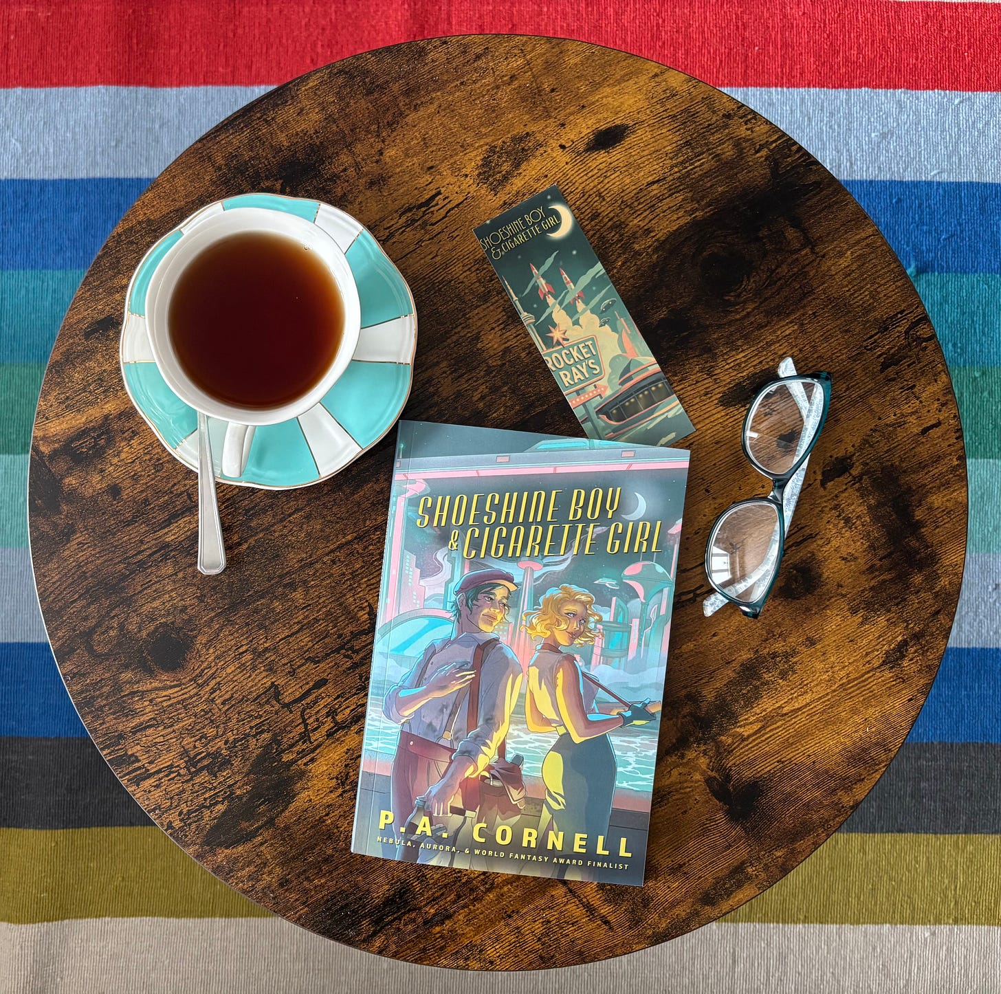 Image shows a small wooden circular table on a colorful striped rug. On the table is a copy of Shoeshine Boy & Cigarette Girl by P.A. Cornell, surrounded by a cup of tea, a bookmark, and a pair of reading glasses.