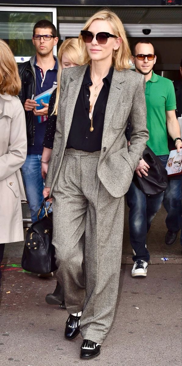 This may contain: a woman in grey suit and black shirt walking down the street with people behind her