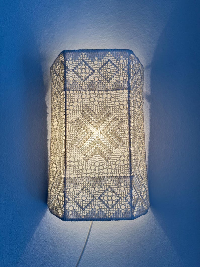 May include: A white, rectangular wall lamp with a detailed, openwork design. The lamp's shade is made of a crocheted material, featuring geometric patterns and a central starburst design. The lamp is mounted on a blue wall, with a white cord extending downwards. May include: A white, rectangular wall lamp with a detailed, openwork design. The lamp's shade is made of a crocheted material, featuring geometric patterns and a central starburst design. The lamp is mounted on a blue wall, with a white cord extending downwards.