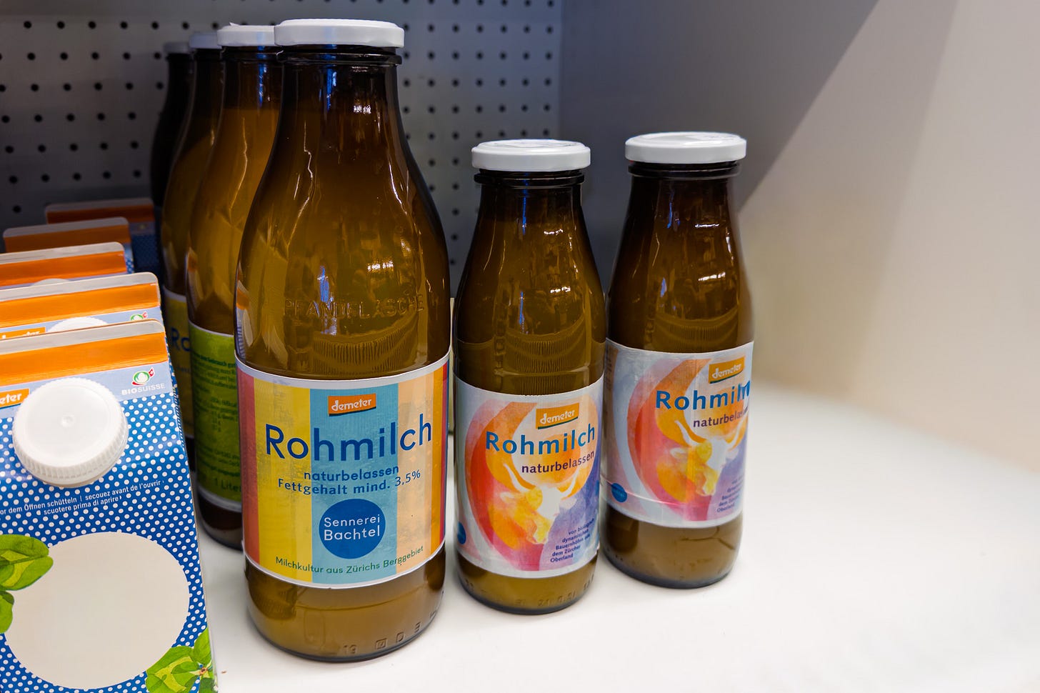 In einem Kühlregal stehen braune Glasflaschen mit weissem Deckel, gefüllt mit Demeter-zertifizierter Rohmilch. Die vordere grosse Ein-Liter-Flasche trägt ein bunt gestreiftes Etikett mit der Aufschrift «Rohmilch naturbelassen, Fettgehalt mind. 3,5 %, Sennerei Bachtel – Milchkultur aus Zürichs Berggebiet». Daneben zwei kleinere Halbliter-Flaschen mit Rohmilch. In einem Kühlregal stehen braune Glasflaschen mit weissem Deckel, gefüllt mit Demeter-zertifizierter Rohmilch. Die vordere grosse Ein-Liter-Flasche trägt ein bunt gestreiftes Etikett mit der Aufschrift «Rohmilch naturbelassen, Fettgehalt mind. 3,5 %, Sennerei Bachtel – Milchkultur aus Zürichs Berggebiet». Daneben zwei kleinere Halbliter-Flaschen mit Rohmilch.
