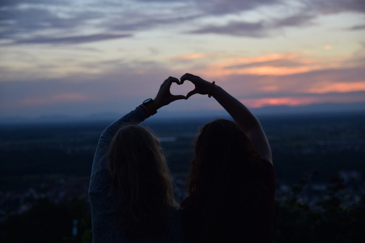 Image of two women seen from the back, looking at a sunset and one arm each raised with their hands forming a heart against the sky.