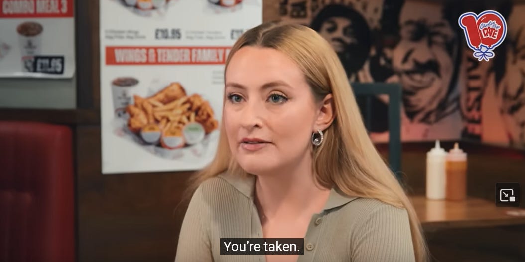 A imagem mostra Amelia dos ombros para cima. Ela é uma mulher branca e loira, com olhos azuis. O fundo da imagem é uma loja fast food de frango, com anúncios de combos de frango frito e batata frita. Ela olha para fora da imagem, triste. A legenda do que ela estava falando no vídeo: You're taken.