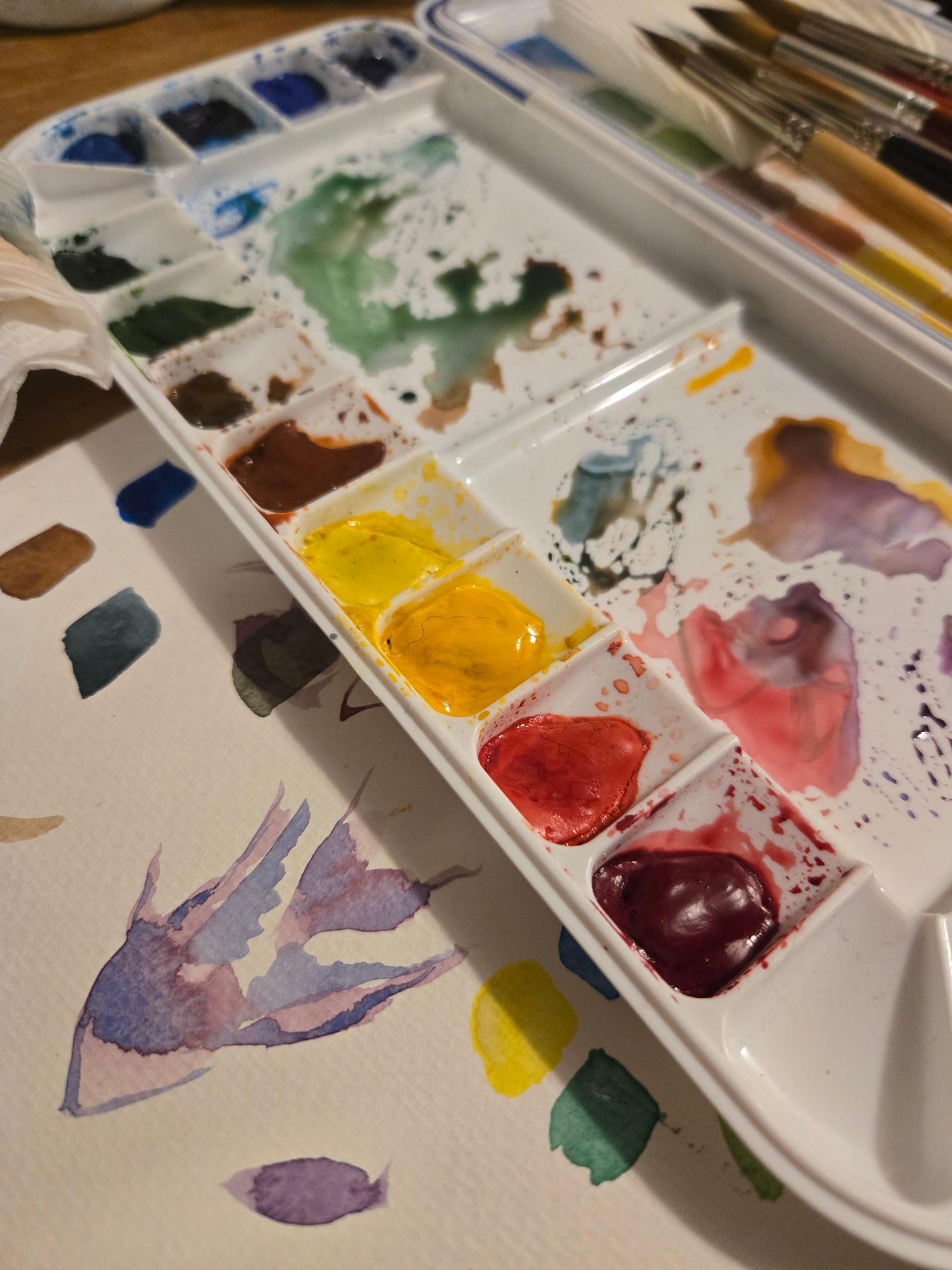 Artist’s watercolor palette with bright pigments and brushes, featuring a mix of yellow, red, blue, and green paints beside a flower sketch.