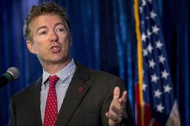 Senator Rand Paul speaks at a microphone in front of an American flag, gesturing with one hand and wearing a red tie. 