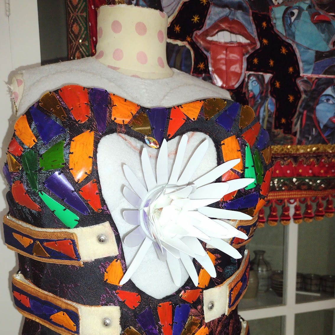A jewel toned mosaic bodice with a white flower bursting forth from the chest. In the background, a tapestry with multiple images of a blue skinned, three eyed goddess with a bright red tongue.