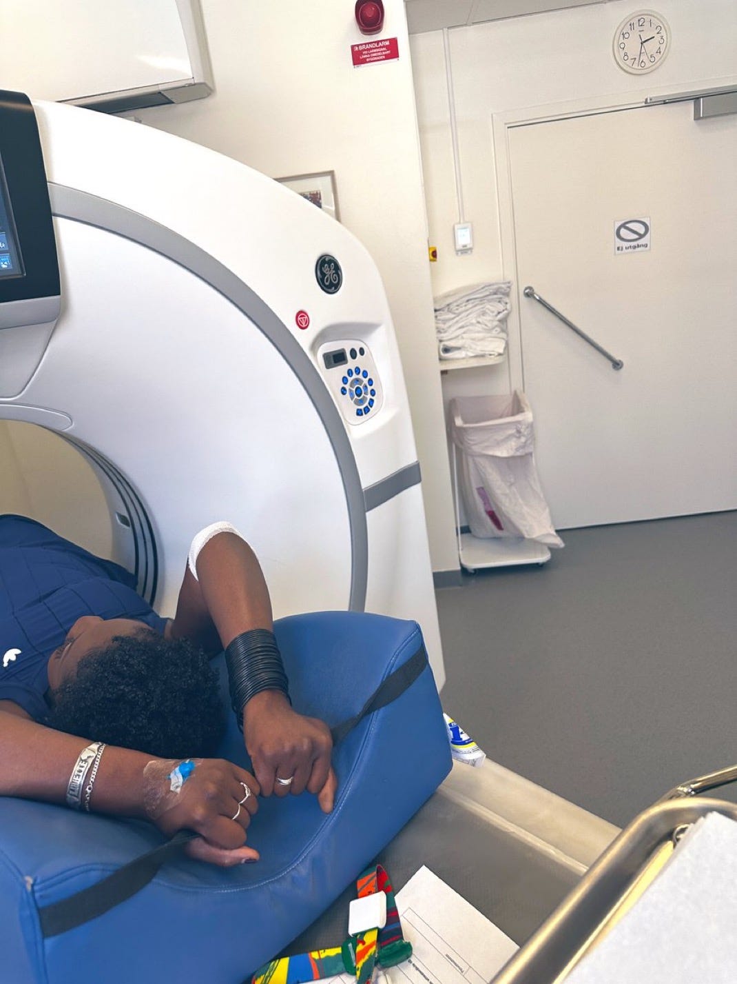 lovette jallow lies on a hospital CT scan table with her arms resting on a blue cushion, IV inserted in one hand and medical wristbands visible. The setting is a sterile hospital room, with folded linens, a clock showing 1:45, and a visible medical machine marked GE. Kente-colored straps and a patient document rest nearby. lovette jallow lies on a hospital CT scan table with her arms resting on a blue cushion, IV inserted in one hand and medical wristbands visible. The setting is a sterile hospital room, with folded linens, a clock showing 1:45, and a visible medical machine marked GE. Kente-colored straps and a patient document rest nearby.