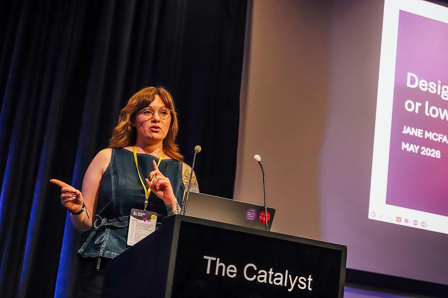 Ellen is standing on stage, mid-speaking and pointing off camera. She has light brown hair and glasses, and is wearing a denim top. She is wearing a yellow lanyard. Behind her is the edge of a slide about dyscalculia.