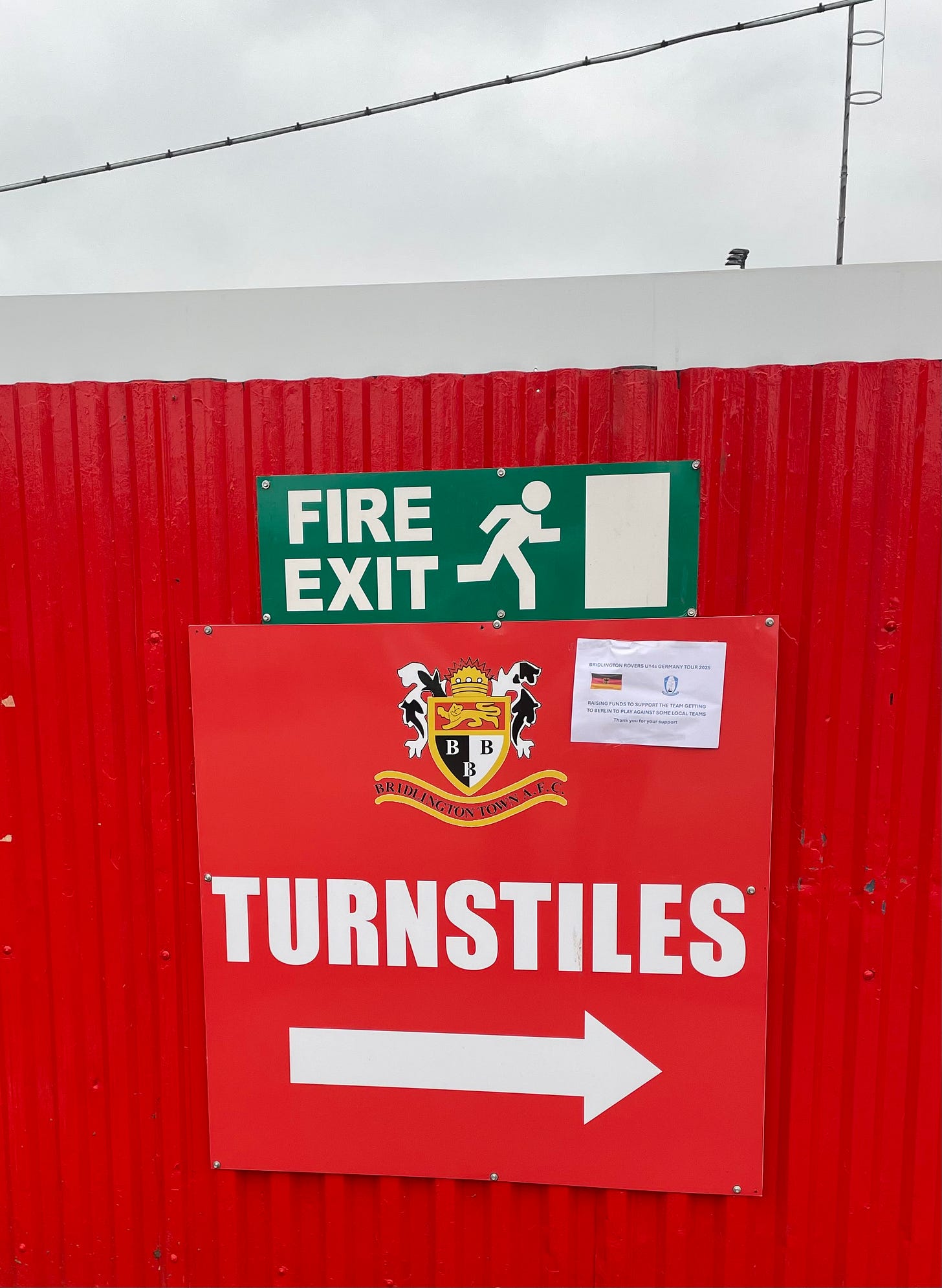 A bright red metal fence with a red sign reading 'Turnstiles' and the Bridlington crest. Above it is a fire exit sign.