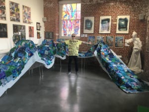 Clarke poses with open arms while standing in the middle of huge sea-like tapestry draped over tables behind him. Clarke poses with open arms while standing in the middle of huge sea-like tapestry draped over tables behind him.