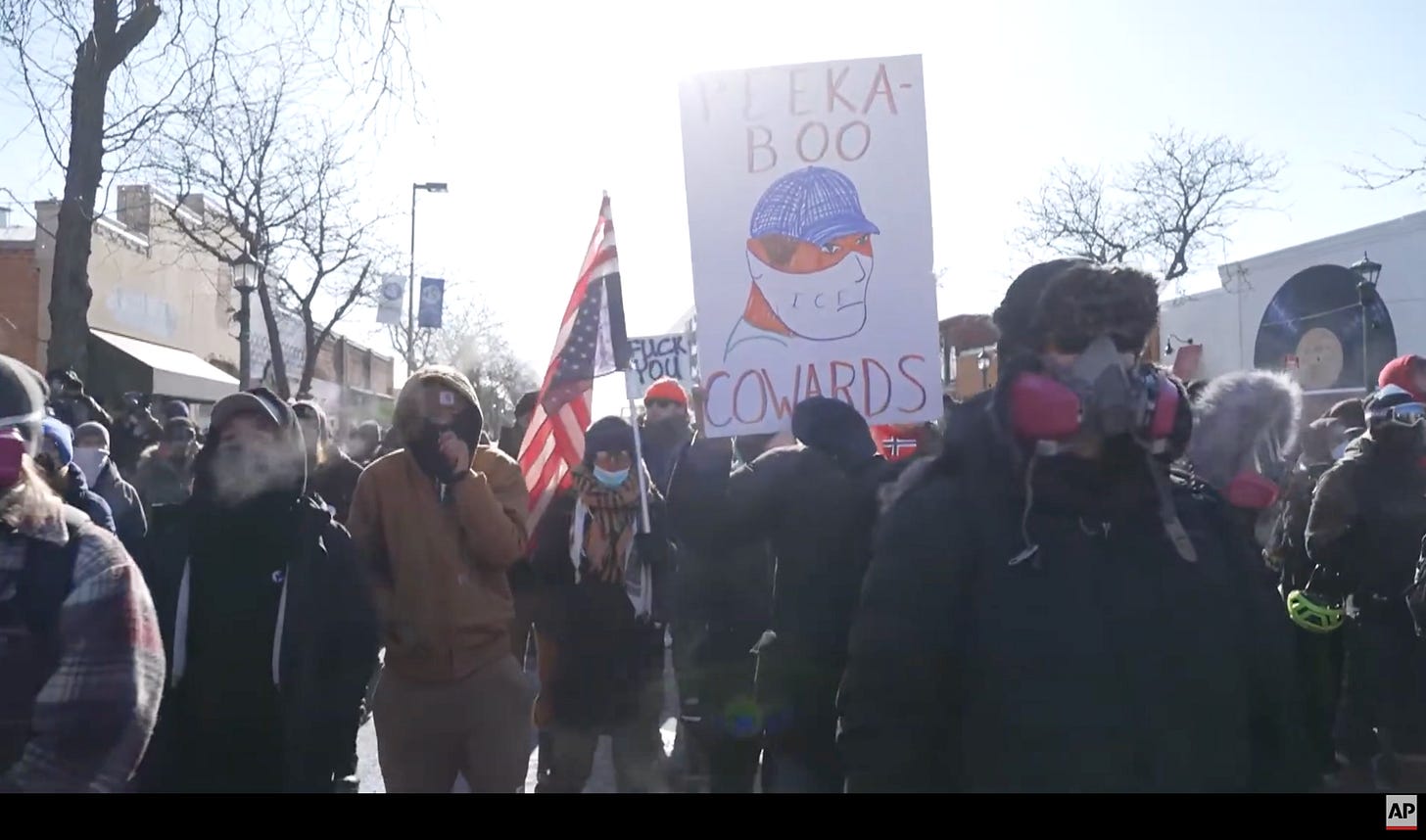 Minneapolis community members in winter clothes, hats, and protective gear are gathered chanting and holding signs, facing federal agents Minneapolis community members in winter clothes, hats, and protective gear are gathered chanting and holding signs, facing federal agents