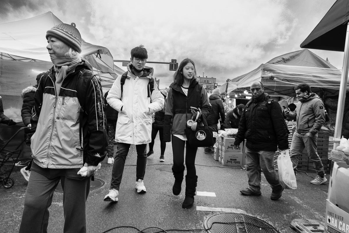 Black and white photo of pedestrians walking through an outdoor Boston market, bundled in winter coats with cloudy skies above. Black and white photo of pedestrians walking through an outdoor Boston market, bundled in winter coats with cloudy skies above.