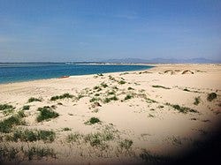 An empty crescent shaped beach, with small patches of grass and a small red boat sits on the shore. The sea appears to the left of the image and mountains appear in the background. An empty crescent shaped beach, with small patches of grass and a small red boat sits on the shore. The sea appears to the left of the image and mountains appear in the background.