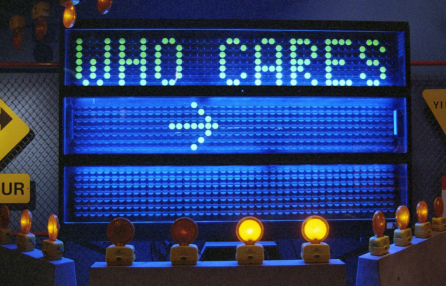 Un cartello elettronico a matrice di punti, illuminato di blu, che visualizza in lettere verdi luminose la frase "WHO CARES" (A CHI IMPORTA). Sotto il testo, un'altra riga di punti luminosi bianchi forma una freccia che punta a destra. Diversi piccoli coni arancioni con luci lampeggianti gialle sono allineati sul bordo inferiore in primo piano. L'illuminazione complessiva è scura e intensa, tipica di un ambiente notturno o interno a tema.