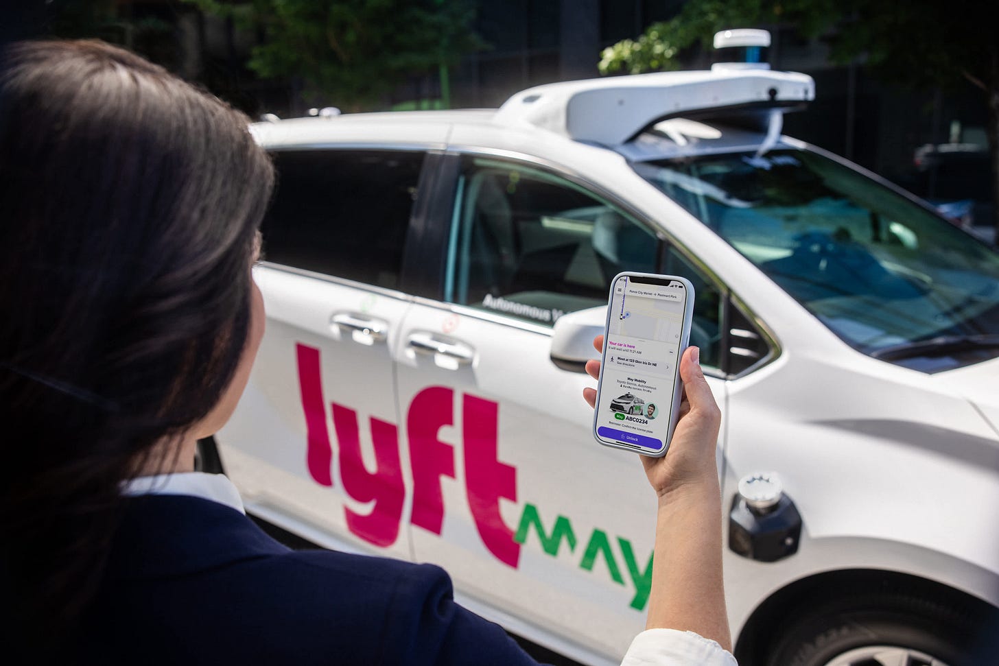 Person holding smartphone with Lyft app open next to white Lyft autonomous vehicle. Person holding smartphone with Lyft app open next to white Lyft autonomous vehicle.