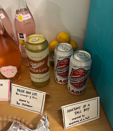 A bowl of pasta salad labeled "Pasta Chives", a Miller High Life for The Holdovers, two tall boys labeled "Anatomy of a Tall Boy," a sign reading "Jeffrey Bites - mac & cheese bites and caramelized onion & feta bites."