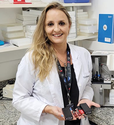 Uma mulher sorridente, de cabelos longos e ondulados, usa um jaleco branco sobre uma blusa preta e um crachá no pescoço. Ela está em um laboratório e segura, com as duas mãos, pequenos dispositivos eletrônicos pretos e vermelhos. Ao fundo, veem-se prateleiras com caixas brancas, equipamentos técnicos sobre a bancada de granito e um fichário azul à direita.