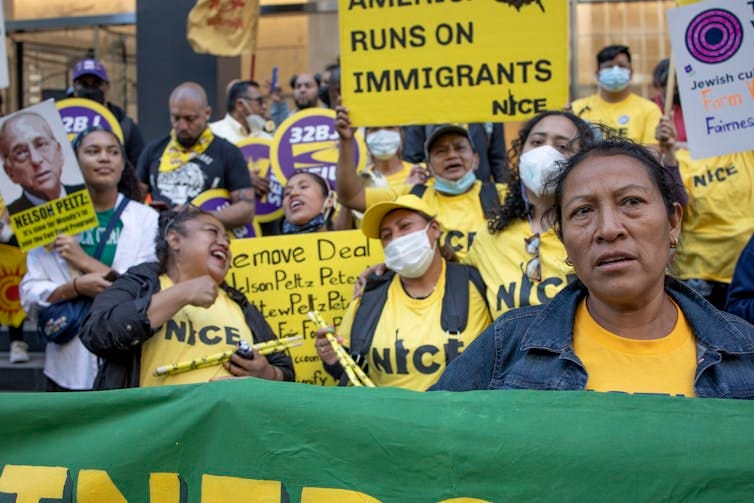 A group of women and men wearing yellow shirts hold signs, one of which says, 'America runs on immigrants.' A group of women and men wearing yellow shirts hold signs, one of which says, 'America runs on immigrants.'