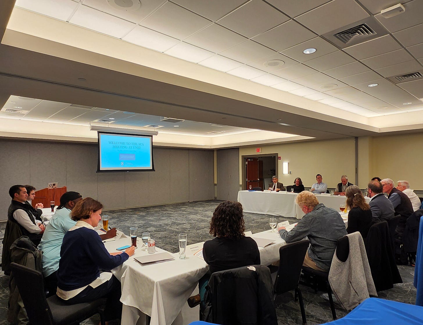 photo of people seated around a U-shaped line of tables, facing a projector screen, in a large conference room photo of people seated around a U-shaped line of tables, facing a projector screen, in a large conference room