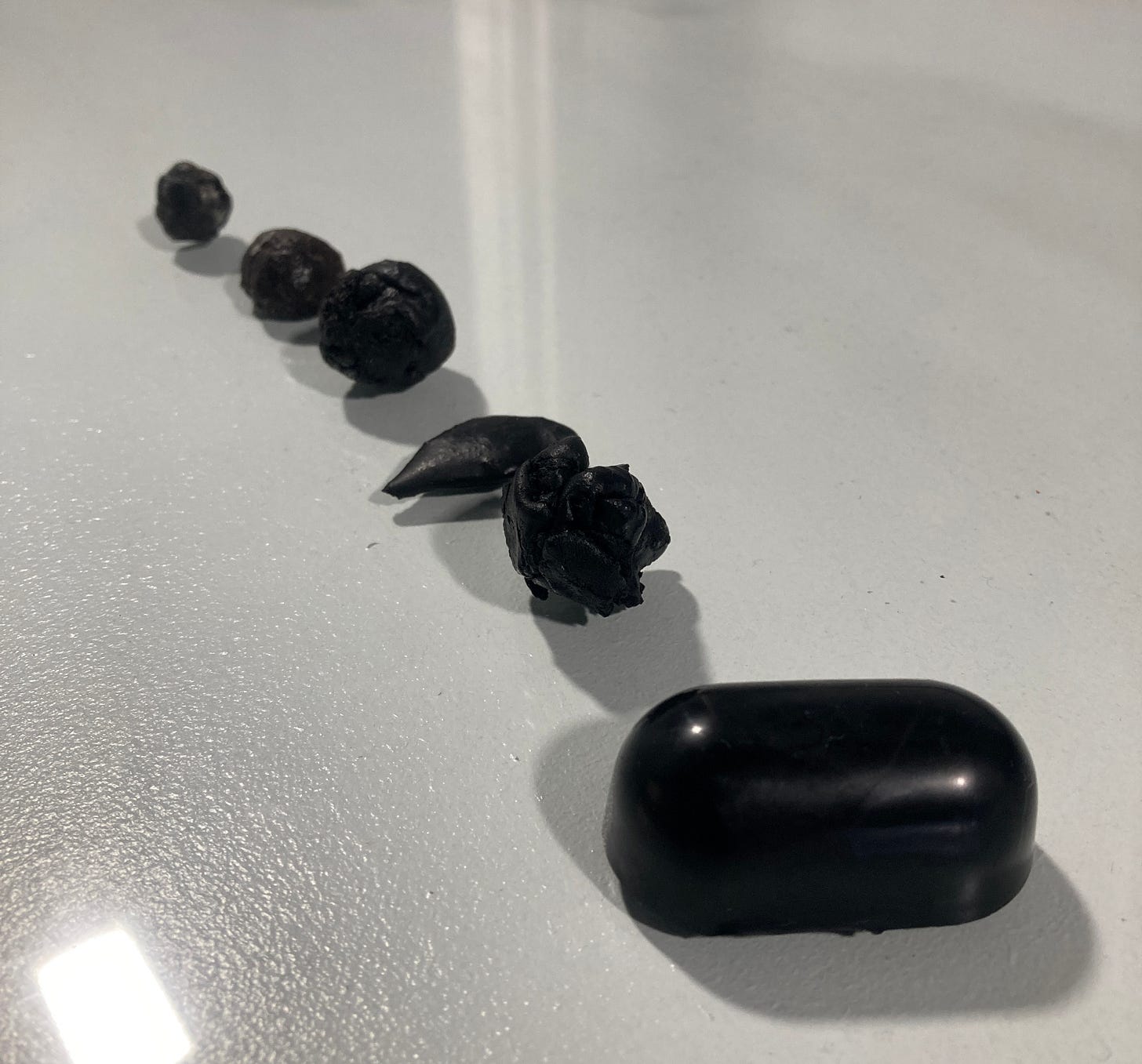 Six versions of ball ground are lined up on a glass slab. The first attempt (back, left), is a small lumpy round ball. Another attempt is smooth and flat like a pancake. The final attempt (front, right) is a large, shiny, smooth pill-shaped block.