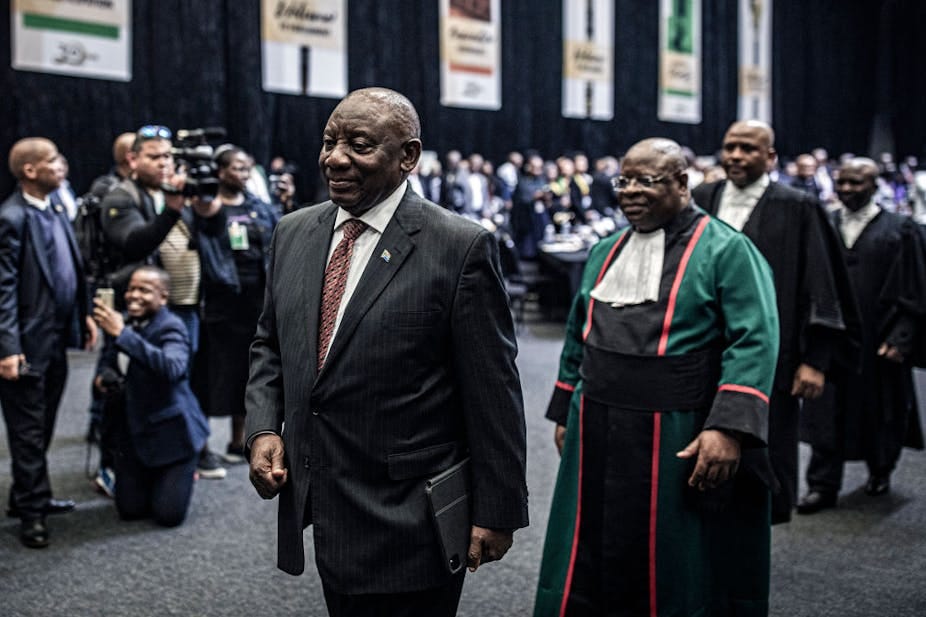 Man in suit and tie walks ahead of a procession of men in gowns, with photographers nearby and more people in the background Man in suit and tie walks ahead of a procession of men in gowns, with photographers nearby and more people in the background