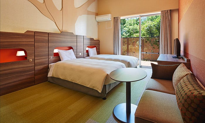 Twin hotel room with wood-paneled headboards and built-in red-lit alcove shelves above each bed. A round table and patterned sofa sit in the foreground on tatami-style green flooring, with a sliding glass door opening onto a balcony with greenery beyond.