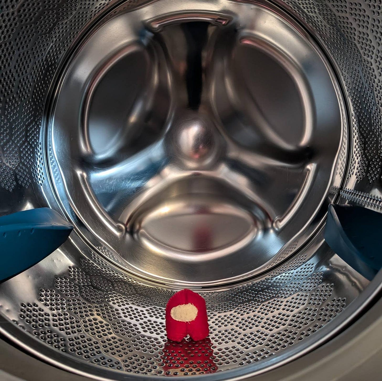 An interior of a washing machine, a small red felt Among Us toy is standing looking out of the drum An interior of a washing machine, a small red felt Among Us toy is standing looking out of the drum