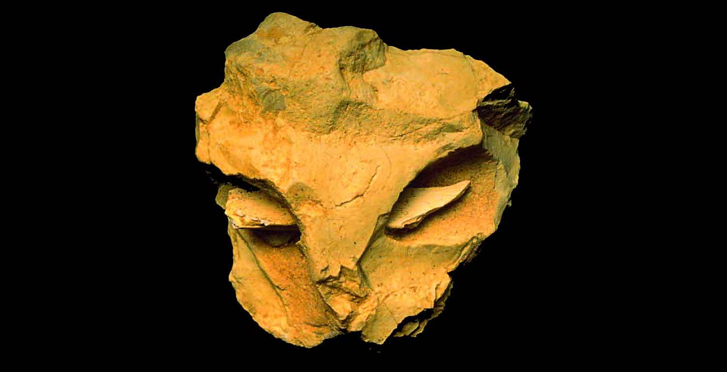 A stone object roughly in the shape of an angular face with a bone splinter highlighting the eyes