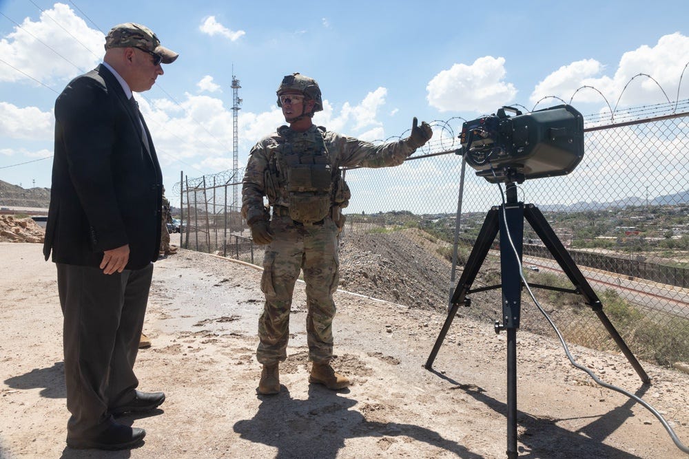 A soldier gestures toward a rectangular acoustic hailer on a tripod beside a border fence while a visitor looks on.