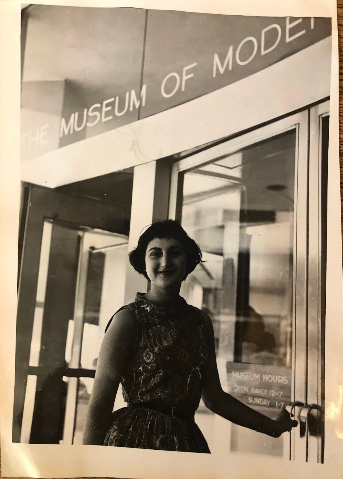 A smiling young woman poses with one hand on the museum entrance door. On the exterior wall above are the partially discernable words, The Museum of Modern