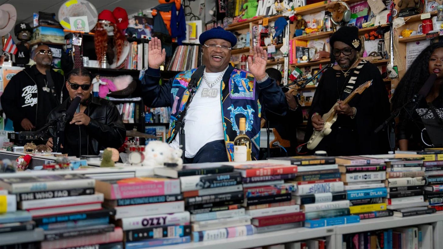 E-40 a happy Black man wearing a colorful jacket, glasses and a blue beanie performs at NPR Tiny Desk surrounded by books and his band.