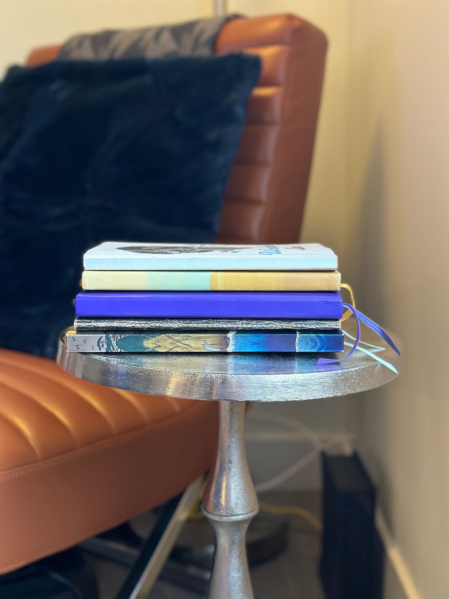 On a silver side table sits a stack of five notebacks. There are assorted colors and sizes. In a blurry background there is a rust-colored leather chair with a blue pillow on it and blanket throws across the back. On a silver side table sits a stack of five notebacks. There are assorted colors and sizes. In a blurry background there is a rust-colored leather chair with a blue pillow on it and blanket throws across the back.