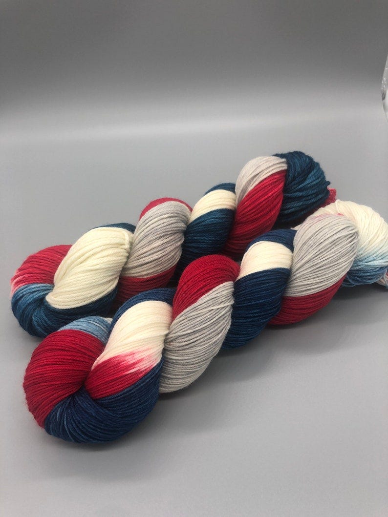 May include: Two skeins of yarn in a red, white, and blue color scheme. The yarn is arranged in a loose coil.