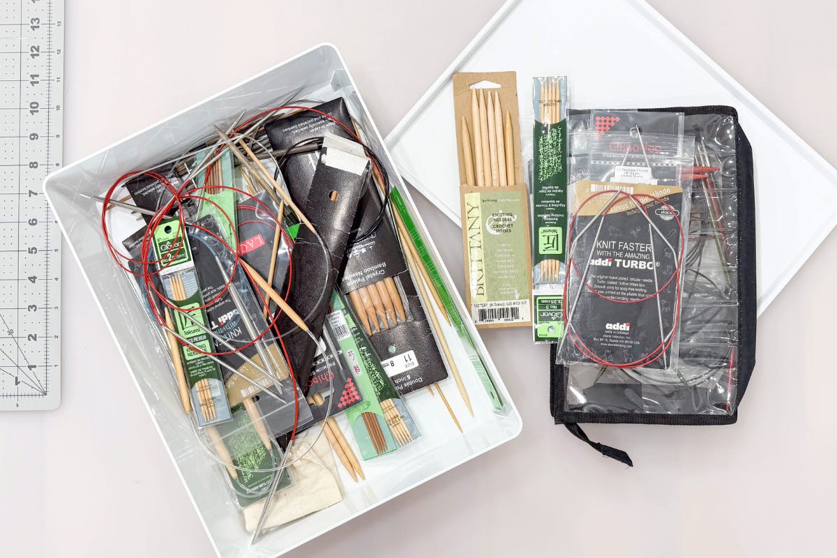 Small white bin holding knitting needles, circular needles, and notions next to cutting mat