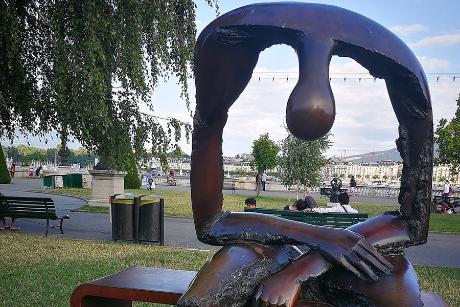 Bronze sculpture of a seated, nude male figure with a large, empty void through the chest and abdomen. The figure leans forward in a posture of sorrow, with the opening revealing the surrounding park behind it.