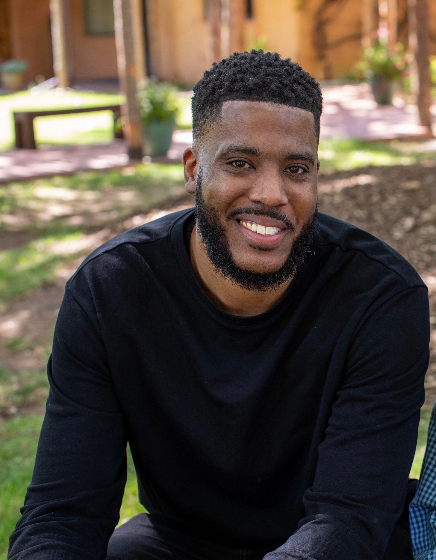 a headshot of a Black man with a beard sitting and smiling, leaning forward, wearing a long-sleeved black shirt, against an outdoor blurred background