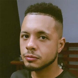 A imagem mostra um retrato em close de uma pessoa voltada para a câmera, com expressão neutra e olhar direto. Ela tem cabelo curto, com as laterais raspadas e o topo um pouco mais volumoso, além de barba e bigode aparados.Veste uma camiseta escura. Ao fundo, aparece um ambiente interno desfocado, com uma parede clara e parte de uma cadeira de madeira. A iluminação é suave e uniforme, destacando o rosto. A foto tem enquadramento dos ombros para cima, semelhante ao de uma foto de perfil.