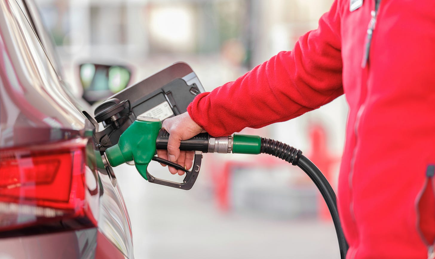 A close-up photograph showing a gas station attendant in a red jacket holding a green fuel nozzle to refuel a car. The image serves as a visual for the news context surrounding Indonesia's state energy company Pertamina raising non-subsidized Pertamax fuel prices by 32.1% to Rp16,250 per liter, sparking middle-class economic concerns and student protests in Jakarta.