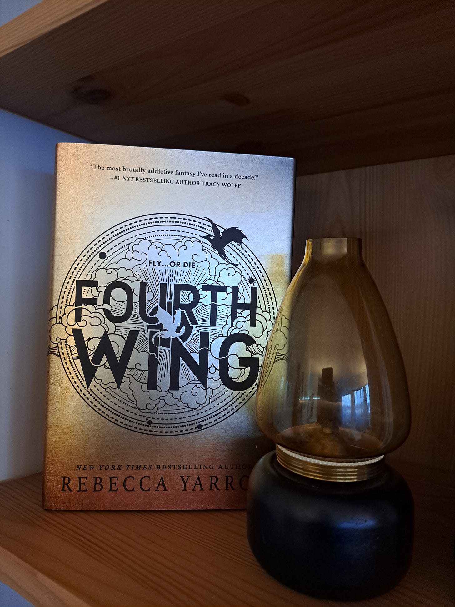 The book Fourth Wing sits on a bookshelf next to a vintage oil lamp