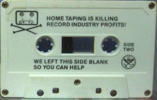 A white cassette tape with two blue wheels and a black window. Text on the tape reads "HOME TAPING IS KILLING RECORD INDUSTRY PROFITS!" and "WE LEFT THIS SIDE BLANK SO YOU CAN HELP." A skull and crossbones symbol is above the first text. The word "SIDE TWO" is printed on the right side.